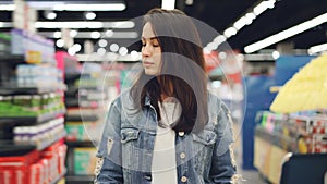 Attractive lady is going through aisle in hypermarket with shopping cart looking at shelves with bright goods. Shopping