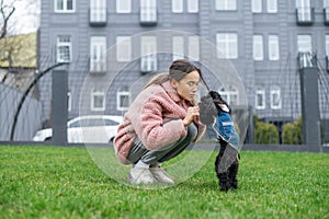 Attractive lady playing with her dog sitting on the lawn against the background of architecture, wearing a pink coat. Cute woman