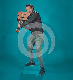 attractive latin man in rock star jacket, on blue background,taking the acoustic guitar like a baseball bat