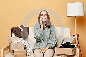Attractive laughing woman sitting on sofa among boxes with clothing against beige wall talking on smartphone with friend saying