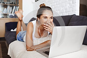 Attractive laughing young girl lying on sofa at living room with laptop.