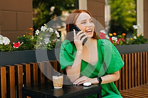 Attractive laughing young woman talking by mobile phone, sitting at table with coffee cup in outdoor cafe terrace in