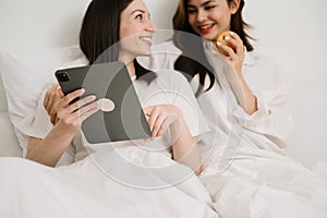 Attractive lesbian couple use mobile phone, tablet watch movie on bed. Beautiful sibling in pajamas lying down with blanket in