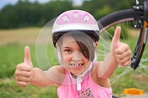 Attractive little cyclist giving thumbs up. Happy joyful little girl with bicycle on the background gesturing thumb up