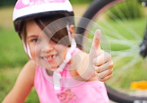 Attractive little cyclist giving thumbs up. Happy joyful little girl with bicycle on the background gesturing thumb up