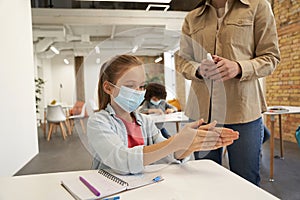 Attractive little school girl wearing protective mask cleaning her hands. Female teacher using an alcohol spray to