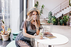 Attractive long-haired girl resting in restaurant and sitting at the table with magazines touching chin with fingers