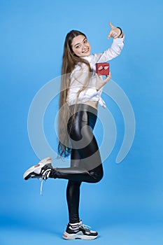 Attractive long-haired lady in leggings pointing at tiny gift box, while shyly bending leg at knee.