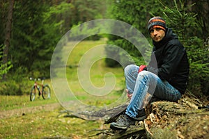 Attractive male cyclist with unshaven in a sports jacket, blue jeans, a buff and trekking boots sits on a tree trunk in the autumn