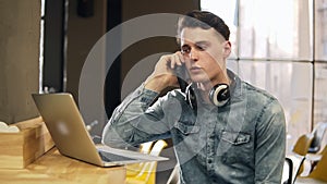 Attractive male in his 20`s in denim shirt with wireless headphones around his neck and laptop beside him, talking with