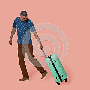 Attractive man carries plastic suitcase on wheels holding it by telescopic handle. Young Caucasian traveler in casual