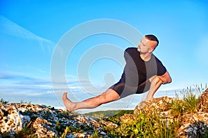 Attractive man doing yoga on the stone against bright blue sky with clouds.