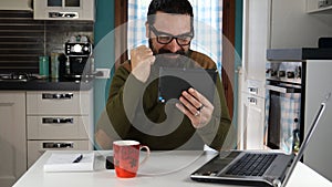 Attractive man with glasses smiling holding tablet and making fist in victory. Sitting at desk in home.