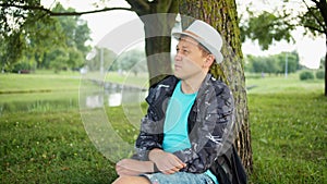 Attractive man in hat resting under a tree in a city park, tracking camera
