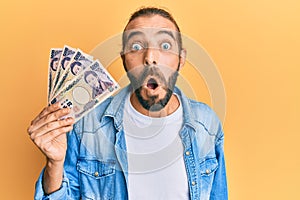 Attractive man with long hair and beard holding 5000 japanese yen banknotes scared and amazed with open mouth for surprise,