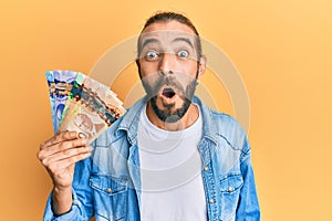 Attractive man with long hair and beard holding canadian dollars scared and amazed with open mouth for surprise, disbelief face