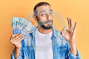 Attractive man with long hair and beard holding south african 100 rand banknotes doing ok sign with fingers, smiling friendly
