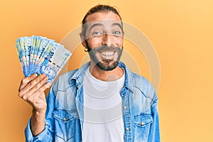 Attractive man with long hair and beard holding south african 100 rand banknotes looking positive and happy standing and smiling