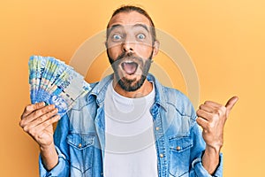Attractive man with long hair and beard holding south african 100 rand banknotes pointing thumb up to the side smiling happy with