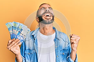 Attractive man with long hair and beard holding south african 100 rand banknotes screaming proud, celebrating victory and success