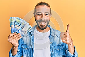 Attractive man with long hair and beard holding south african 100 rand banknotes smiling happy and positive, thumb up doing