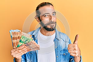 Attractive man with long hair and beard holding south african rand banknotes smiling happy and positive, thumb up doing excellent