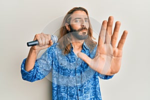 Attractive man with long hair and beard singing song using microphone with open hand doing stop sign with serious and confident