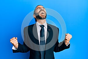 Attractive man with long hair and beard wearing business suit and tie very happy and excited doing winner gesture with arms