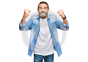 Attractive man with long hair and beard wearing casual denim jacket angry and mad raising fists frustrated and furious while