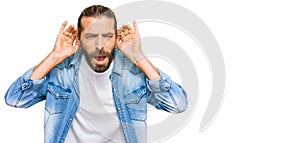 Attractive man with long hair and beard wearing casual denim jacket trying to hear both hands on ear gesture, curious for gossip