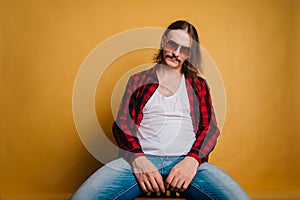 Attractive man with long hair and a mustache in a red 80s disco plaid shirt, sitting on a chair against a yellow background. Retro