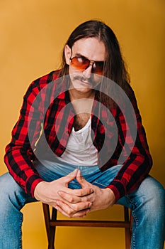 Attractive man with long hair and a mustache in a red 80s disco plaid shirt, sitting on a chair against a yellow background. Retro