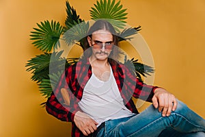 An attractive man with long hair and a mustache in a red plaid 80s disco shirt sits on a chair against a yellow background. Guy in