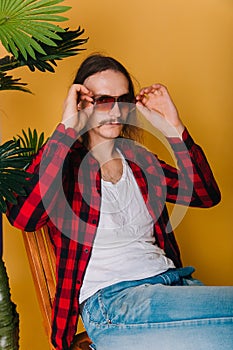 An attractive man with long hair and a mustache in a red plaid 80s disco shirt sits on a chair against a yellow background. Guy in