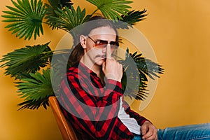 An attractive man with long hair and a mustache in a red plaid 80s disco shirt sits on a chair against a yellow background. Guy in