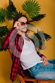 An attractive man with long hair and a mustache in a red plaid 80s disco shirt sits on a chair against a yellow background. Guy in