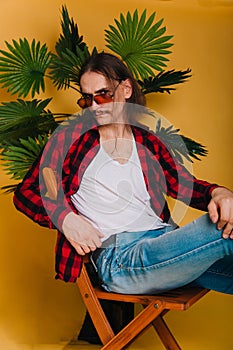 An attractive man with long hair and a mustache in a red plaid 80s disco shirt sits on a chair against a yellow background. Guy in