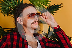 An attractive man with long hair and a mustache in a red plaid 80s disco shirt sits on a chair against a yellow background. Guy in