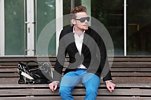 An attractive man sitting down outside wearing jeans and a blazer