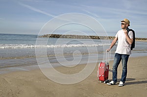 Attractive man traveler in a hat and with a red suitcase stands on the seashore, taking off his jacket, place for text