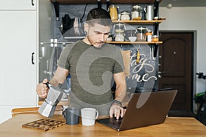 Attractive man uses a laptop and pours coffee in the interior of a home kitchen in the morning.