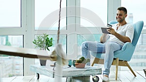 Attractive man using digital tablet sitting in chair at balcony in loft modern apartment