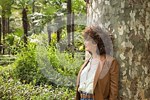 Attractive mature woman with curly brown hair, wearing a brown leather jacket and jeans leaning against a tree trunk, pensive and