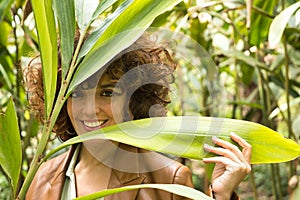Attractive mature woman with curly brown hair, wearing brown leather jacket looking through large green leaves of a plant. Concept