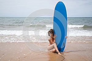 Attractive mature woman with curly hair, sunglasses and bikini, sitting clutching a blue surfboard on the shore of the beach.