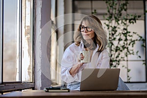 Attractive mid aged woman sitting at window and using her laptop for work