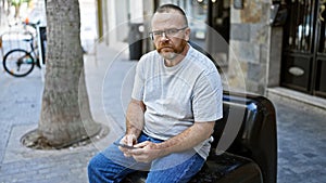 Attractive middle-aged caucasian man, with a serious expression, avidly typing a message on his smartphone while sitting outdoors