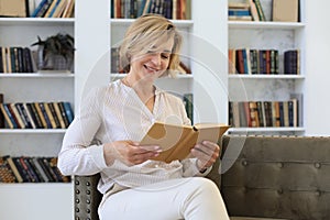 Attractive middle aged woman enjoying reading a book sitting on the sofa in her living room smiling while she reading