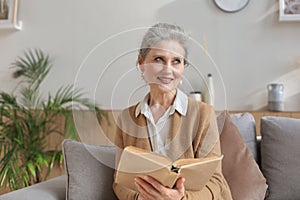 Attractive middle aged woman enjoying reading a book sitting on the sofa in her living room smiling while she reading