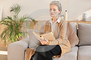 Attractive middle aged woman enjoying reading a book sitting on the sofa in her living room smiling while she reading.